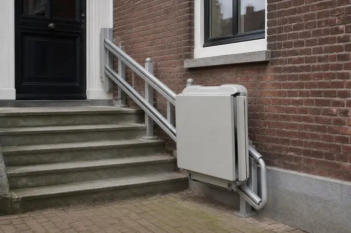 AI-beeld: een opgeklapt plateau van een plateaulift tegen een bakstenen muur, naast vijf stenen treden naar een voordeur, met de rail langs de treden
