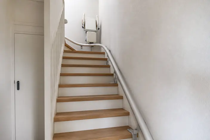 AI-beeld: een traplift met een rail langs een trap met houten treden en een bocht bovenaan, waar de lege stoel staat