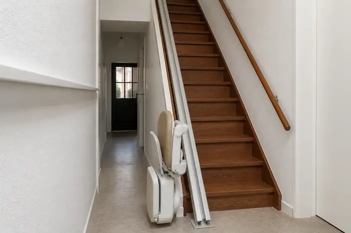 AI-beeld: een lege traplift met opgeklapte zitting, armleuningen en voetenplank onder aan een rechte houten trap in de gang van een rijtjeshuis, met de rail langs de trap omhoog